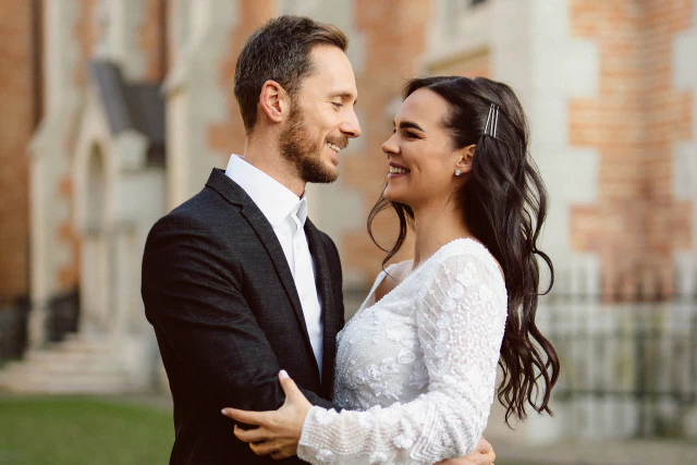 Mosolygó menyasszony és vőlegény egymást átölelve egy épület előtt.