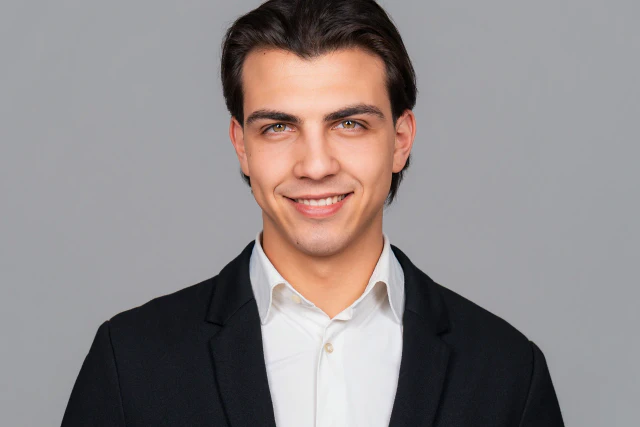 Headshot of a smiling young man wearing a dark jacket and white shirt against a clean grey background