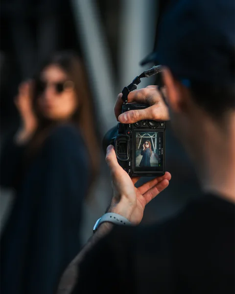 A photographer takes a portrait of a woman wearing sunglasses, with the finished image visible on the camera screen