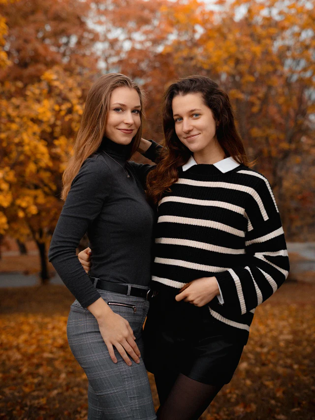 Two young women standing together in an autumn park with orange foliage behind them