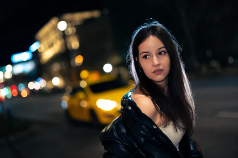 Fekete bőrdzsekis nő éjszakai városi fények és egy sárga taxi előtt.