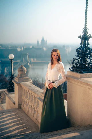 Fehér blúzos nő zöld szoknyában áll egy városi panoráma fölötti lépcsőn.