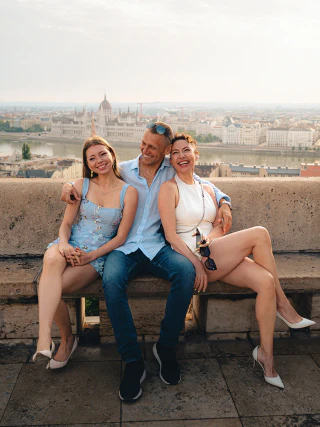 A cheerful family of three sitting on a bench with the Budapest skyline and Parliament behind them