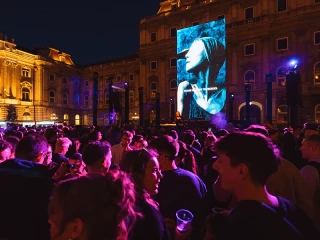 Közönség egy esti szabadtéri koncerten, nagyméretű színpadi kijelző előtt.