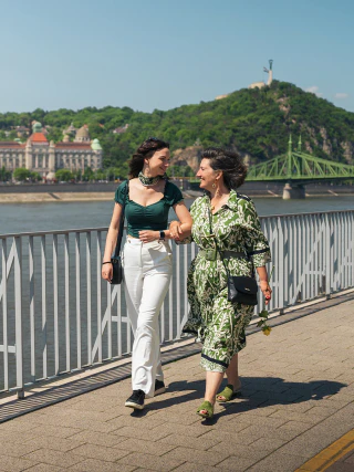A mother and her adult daughter walking beside the Danube with Gellért Hill and Liberty Bridge behind them