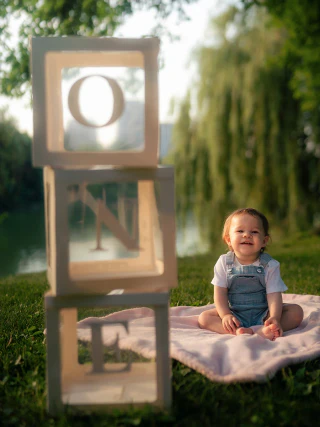 A smiling young child sitting on a blanket beside wooden blocks spelling ONE