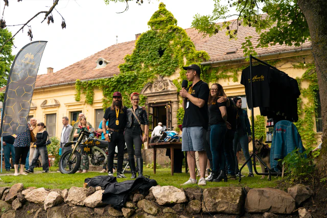 Mikrofont tartó férfi beszél egy szabadtéri rendezvényen, kollégáival és kiállított motorokkal a háttérben.