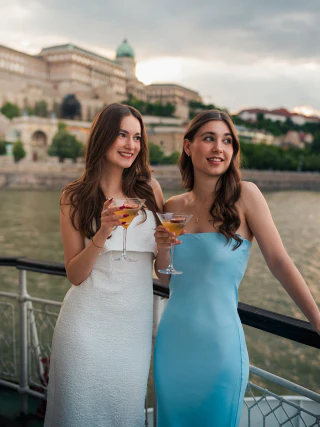 Two young women holding cocktails on the deck of a boat with Buda Castle behind them