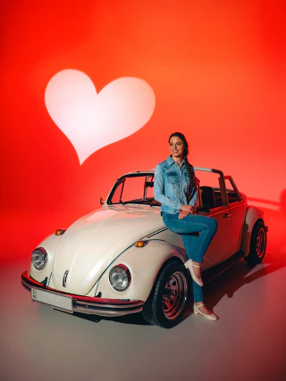 Nő ül egy fehér Volkswagen Bogár kabrió elején piros stúdióháttér előtt