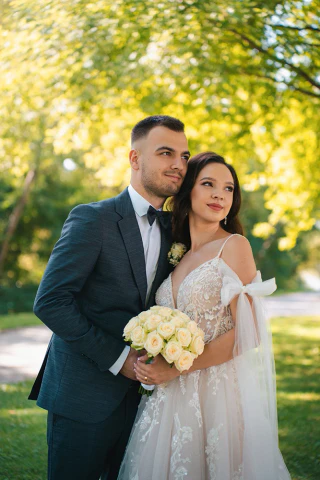 Vőlegény átöleli a menyasszonyt egy napsütötte parkban.