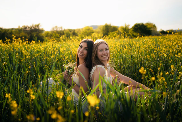 Two young women sitting back to back in a sunny field of yellow flowers while holding small bouquets