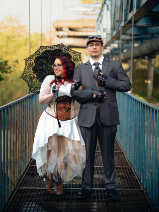 A steampunk-style bride and groom standing on a metal bridge with a lace parasol.