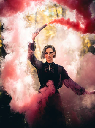 A woman in a dark outfit holding a smoke flare in a cloud of red and pink smoke