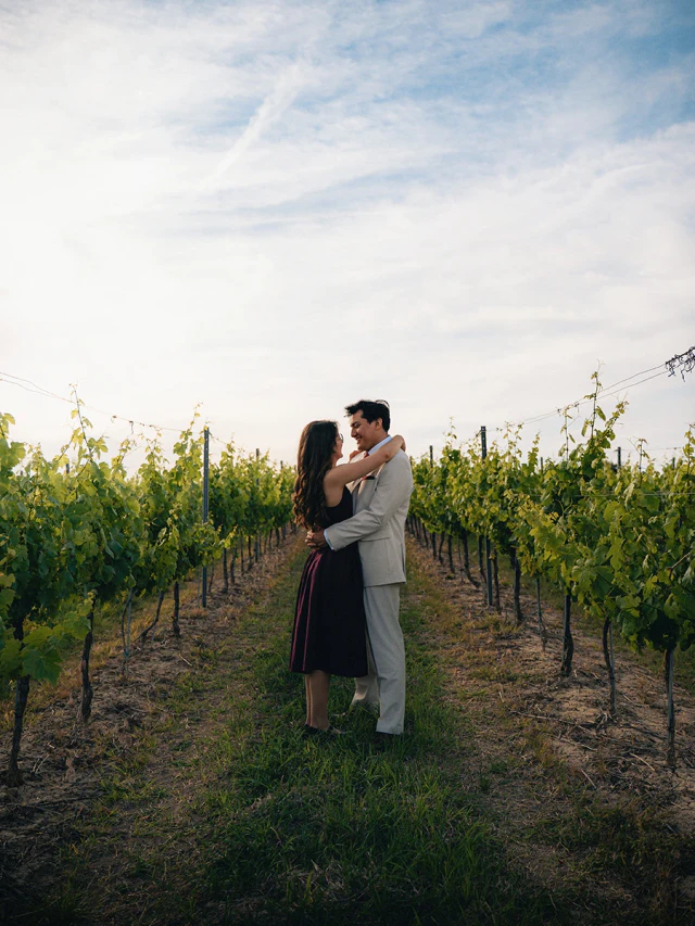 An elegantly dressed couple embracing between rows of vines.