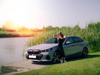 Nő támaszkodik egy ezüstszínű BMW-re egy tóparti helyszínen.