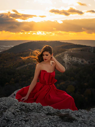 A woman in a red dress sitting on a rock at sunset with mountains in the background