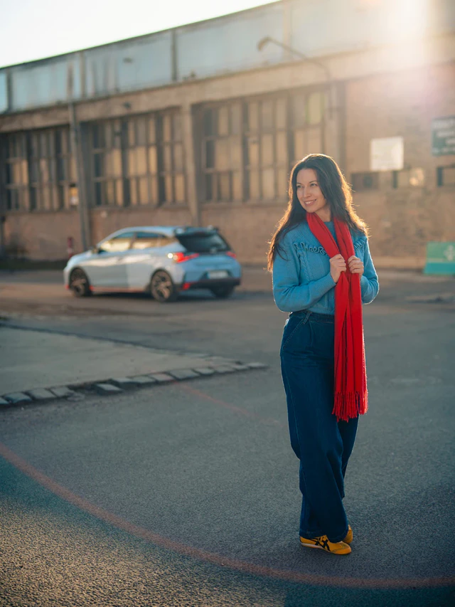Nő autós portré piros sállal, ipari környezetben, háttérben egy világoskék autóval