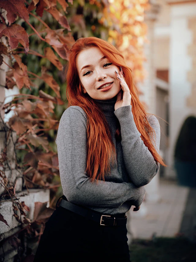 A red-haired woman in a grey roll-neck jumper posing beside a wall covered in autumn leaves