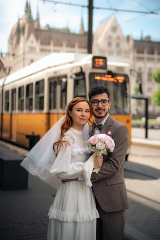 Esküvői pár áll egy sárga villamos előtt, a menyasszony virágcsokorral.