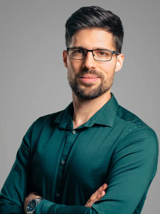 Male headshot wearing glasses and a green shirt with folded arms against a grey background