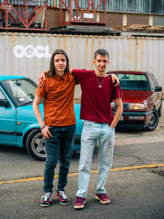Two young men standing beside parked cars with one placing his arm around the other