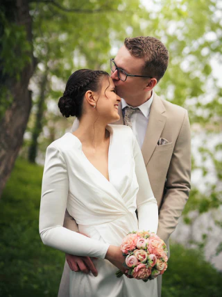 A groom kisses the bride on the forehead as she holds a bouquet in a park.