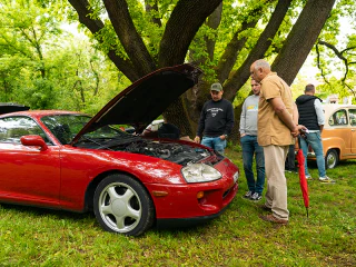 Látogatók egy nyitott motorháztetejű piros sportautót néznek egy szabadtéri autós rendezvényen.