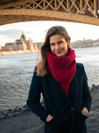 A woman in a dark coat and red scarf standing beside the Danube, with the Hungarian Parliament Building and Margaret Bridge in the background
