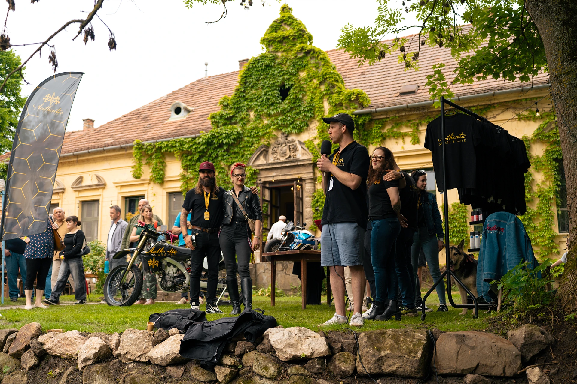 A man speaks into a microphone at an outdoor event with colleagues and motorcycles behind him.