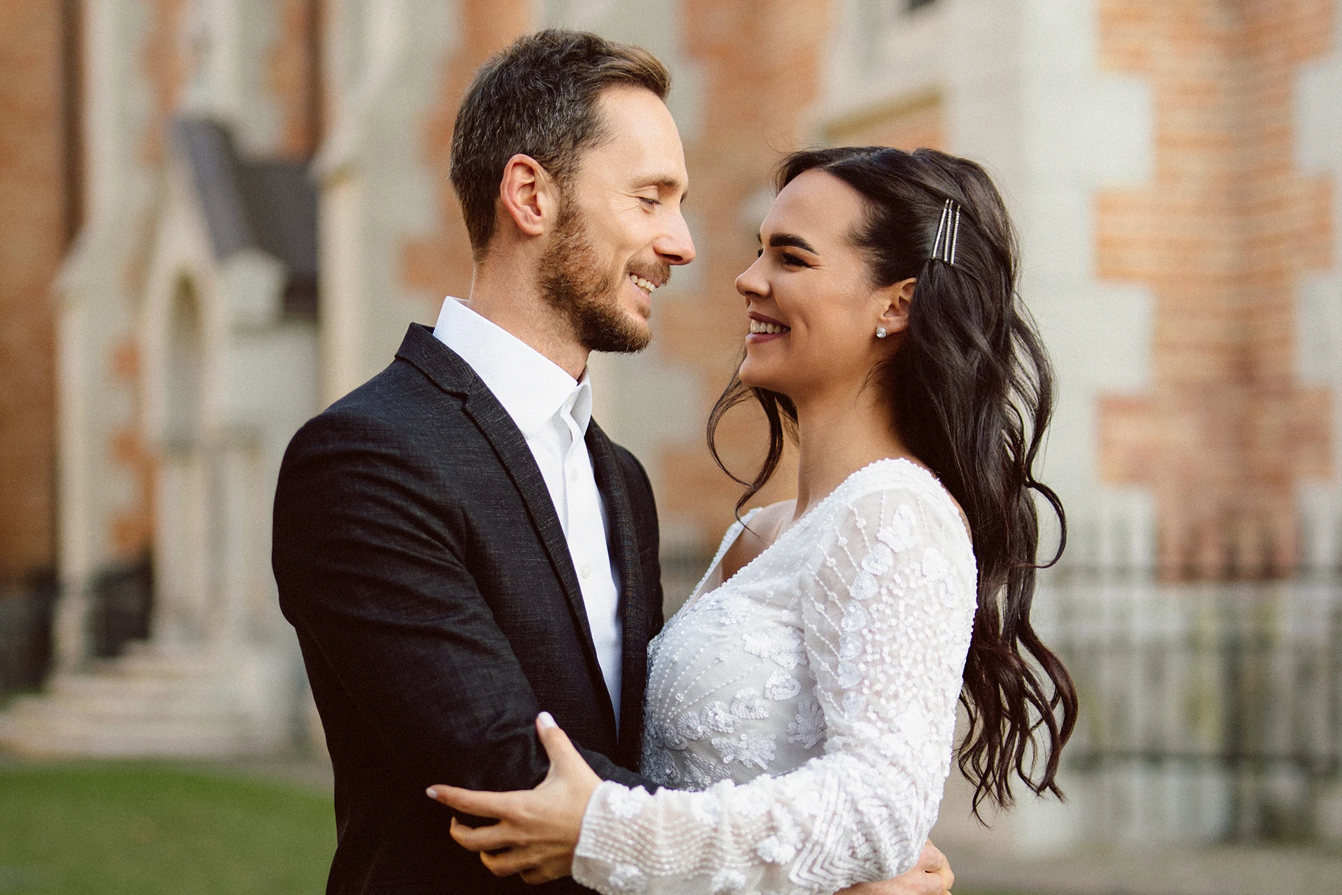 Mosolygó menyasszony és vőlegény egymást átölelve egy épület előtt.