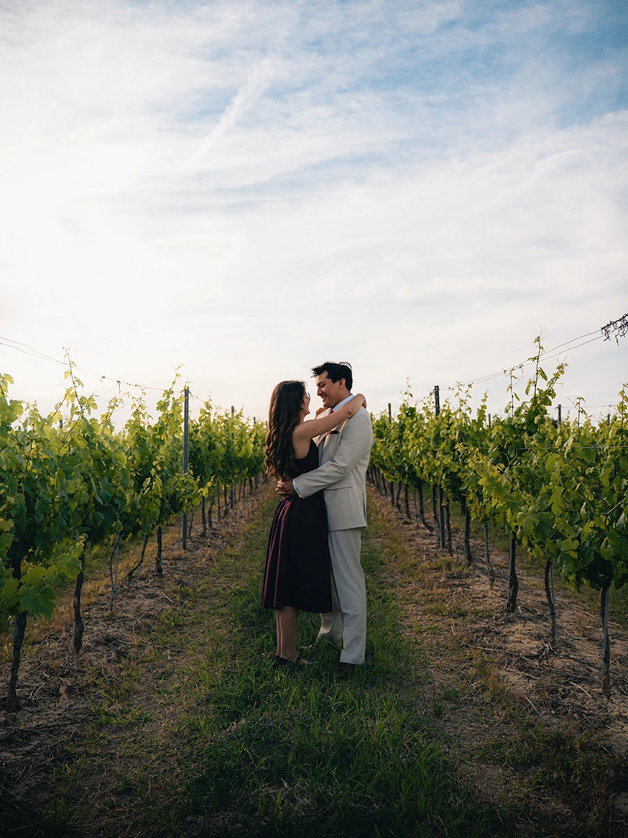 An elegantly dressed couple embracing between rows of vines.