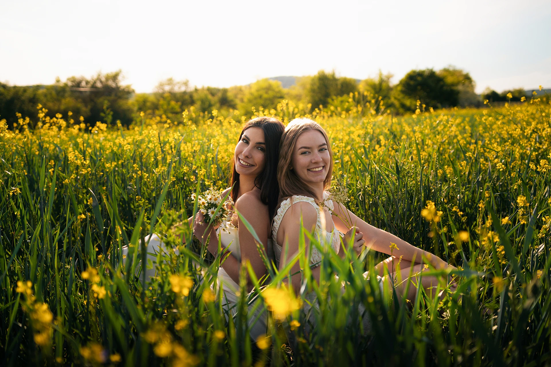 Two young women sitting back to back in a sunny field of yellow flowers while holding small bouquets